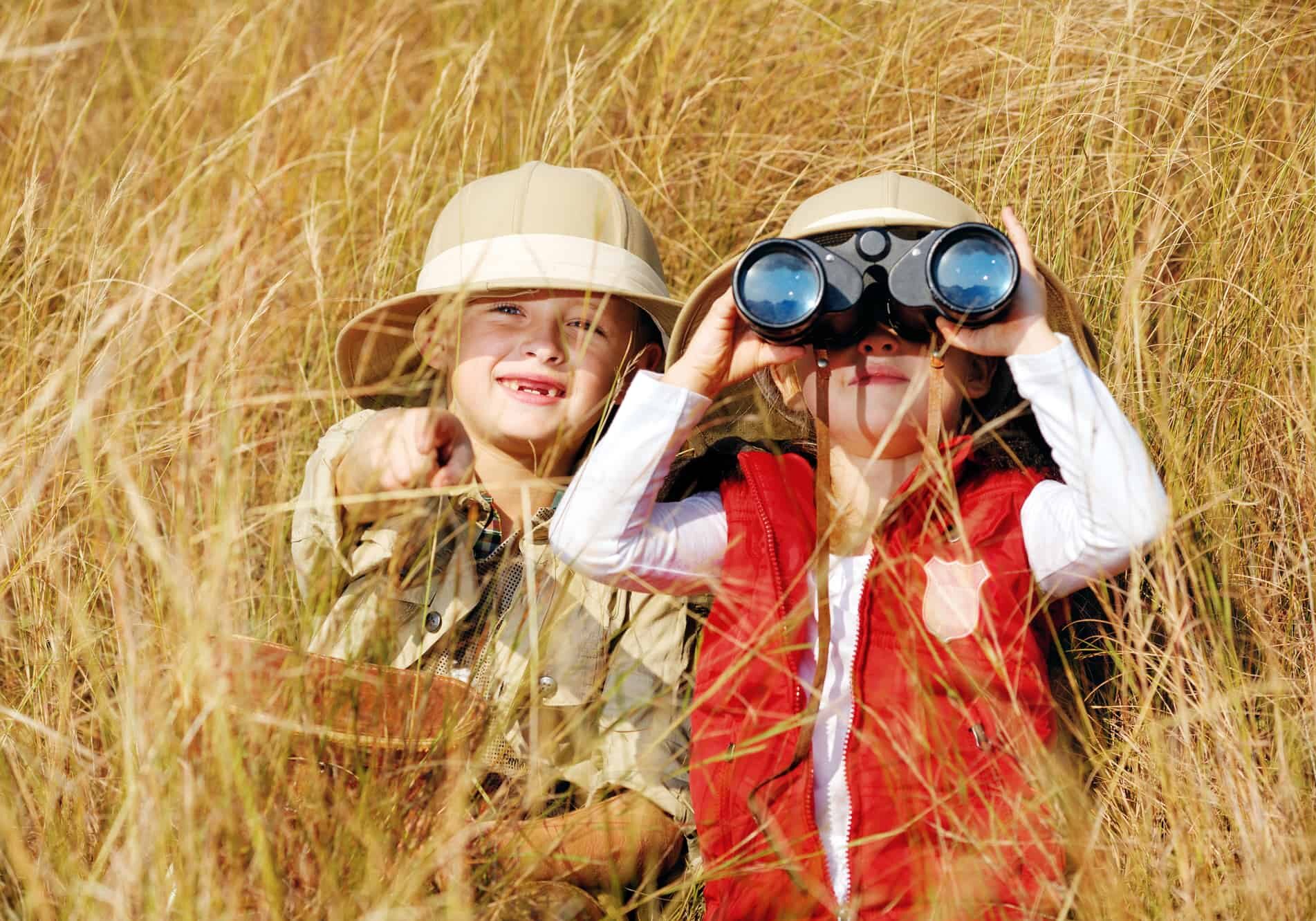 Happy young safari adventure children playing outdoors in the grass with binoculars and exploring together as brother and sister.